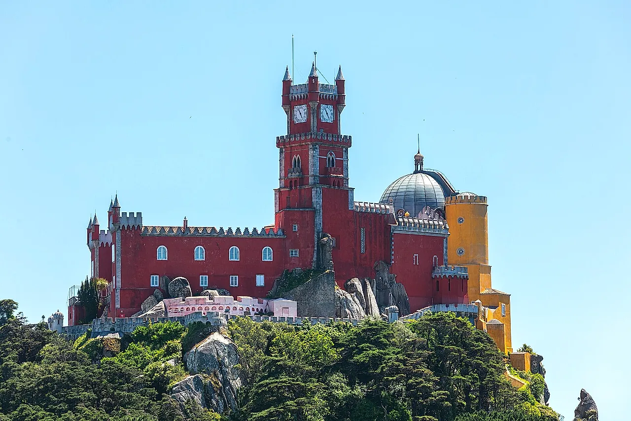 The red clock tower and walls of Pena Palace crowning a wooded hilltop under a clear blue sky