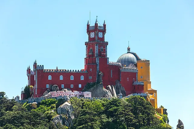 The red clock tower and walls of Pena Palace crowning a wooded hilltop under a clear blue sky